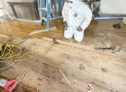 A person in a white hazmat suit kneels on a partially removed wooden floor, revealing the subfloor and scattered tools. The area appears to be under renovation, with plastic sheeting in the background.