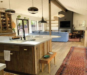 An open-concept kitchen and living room features a rustic wood island, light blue sofa, and warm rugs on hardwood floors. Pendant lights hang above.