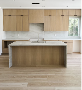 A modern kitchen features light wood cabinets, light stone countertops and backsplash, with a large island and sink. Light wood flooring completes the bright, minimalist space.