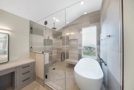A spacious, modern bathroom featuring a large glass shower enclosure, a sleek freestanding tub, and a vanity with drawers. Light-toned, patterned tiles adorn the walls and shower interior.